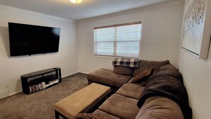 Una sala de estar con un sofá modular, un televisor de pantalla plana montado en la pared, una ventana con persianas y un mueble de entretenimiento negro con libros.