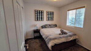 Un dormitorio con una cama, una mesita de noche, dos obras de arte en la pared, una ventana con persianas y suelo alfombrado.