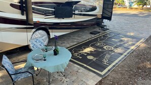 Een camper met een overdekt terras, een tafel met stoelen en een decoratieve mat.