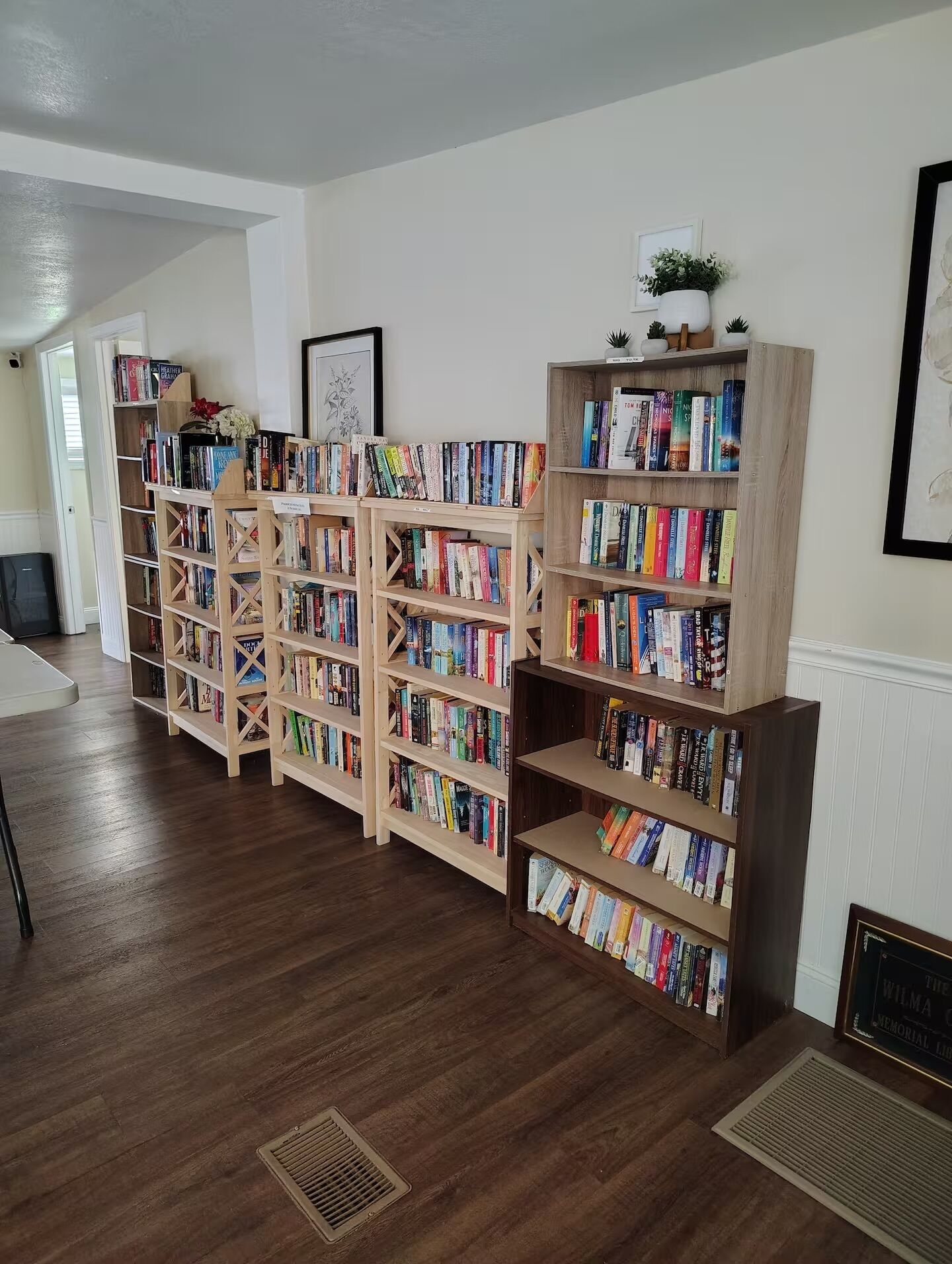 Una biblioteca con varias estanterías de madera llenas de libros, un cuadro en la pared y una rejilla en el suelo.