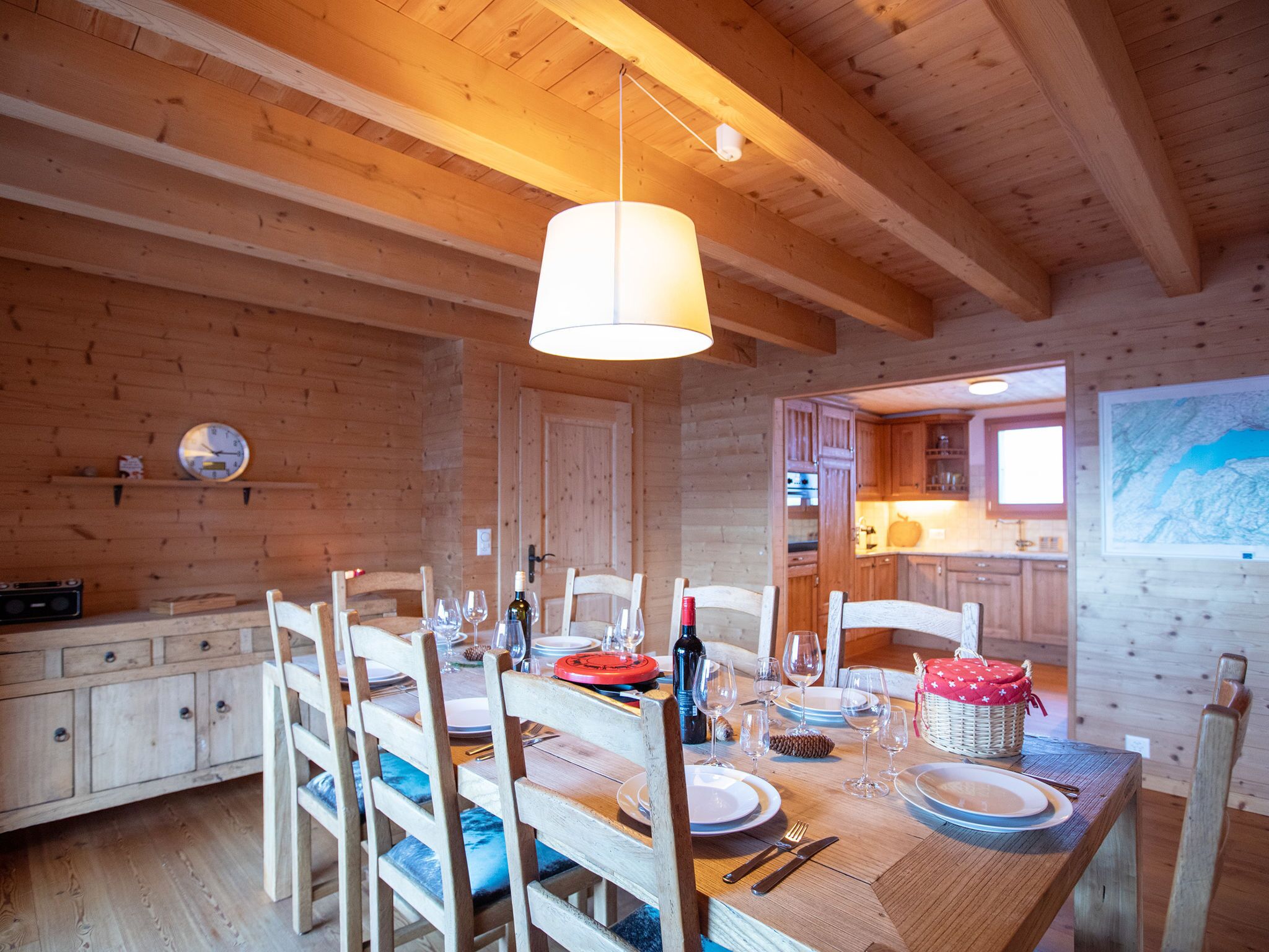Un intérieur en bois avec une table à manger dressée pour quatre personnes, des chaises en bois, une cuisine visible à travers une porte ouverte et un luminaire suspendu.