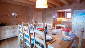 A wooden interior with a dining table set for four, wooden chairs, a kitchen visible through an open door, and a hanging light fixture.