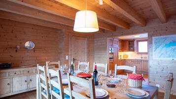 Un intérieur en bois avec une table à manger dressée pour quatre personnes, des chaises en bois, une cuisine visible à travers une porte ouverte et un luminaire suspendu.