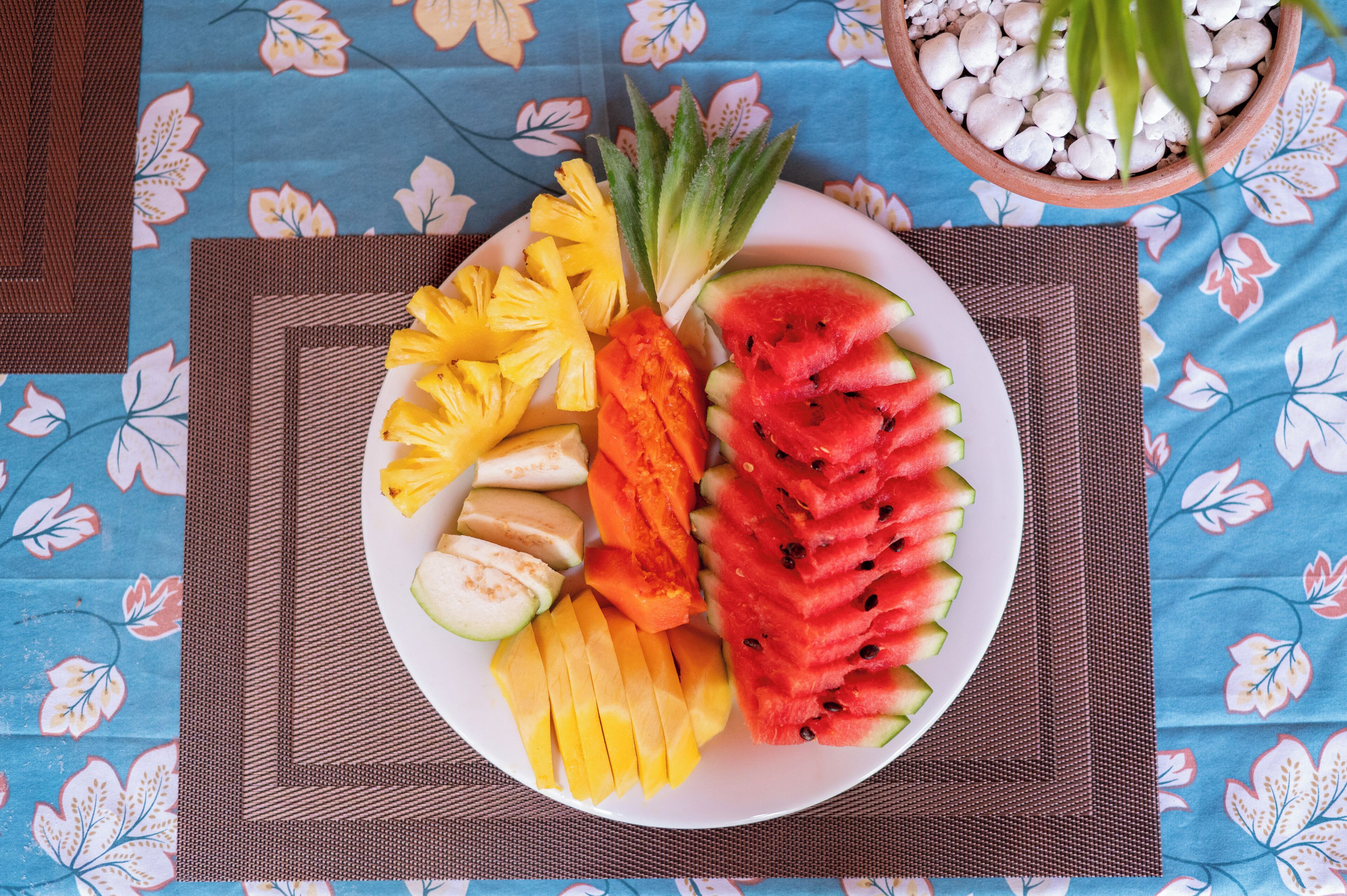 A plate of sliced fruits including pineapple, mango, watermelon, and papaya, placed on a table with a blue floral tablecloth.