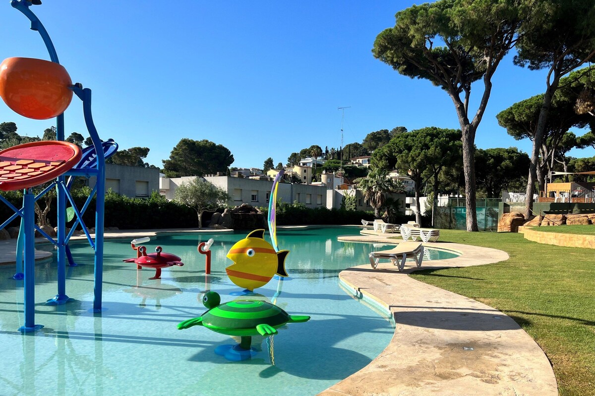 A swimming pool area with colorful play structures, including a fish and a turtle, surrounded by a well-maintained lawn and trees.