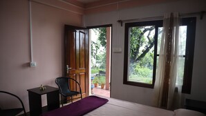 A bedroom with a bed, a chair, a small table, and a window with a view of greenery outside. - The Bicknell's Nest (Kalimpong)
