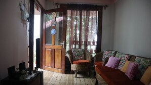 A room with a wooden floor, a wicker sofa with colorful cushions, a wicker chair with a cushion, a desk with a computer and a trash bin, and a door with a window and curtains. - The Bicknell's Nest (Kalimpong)