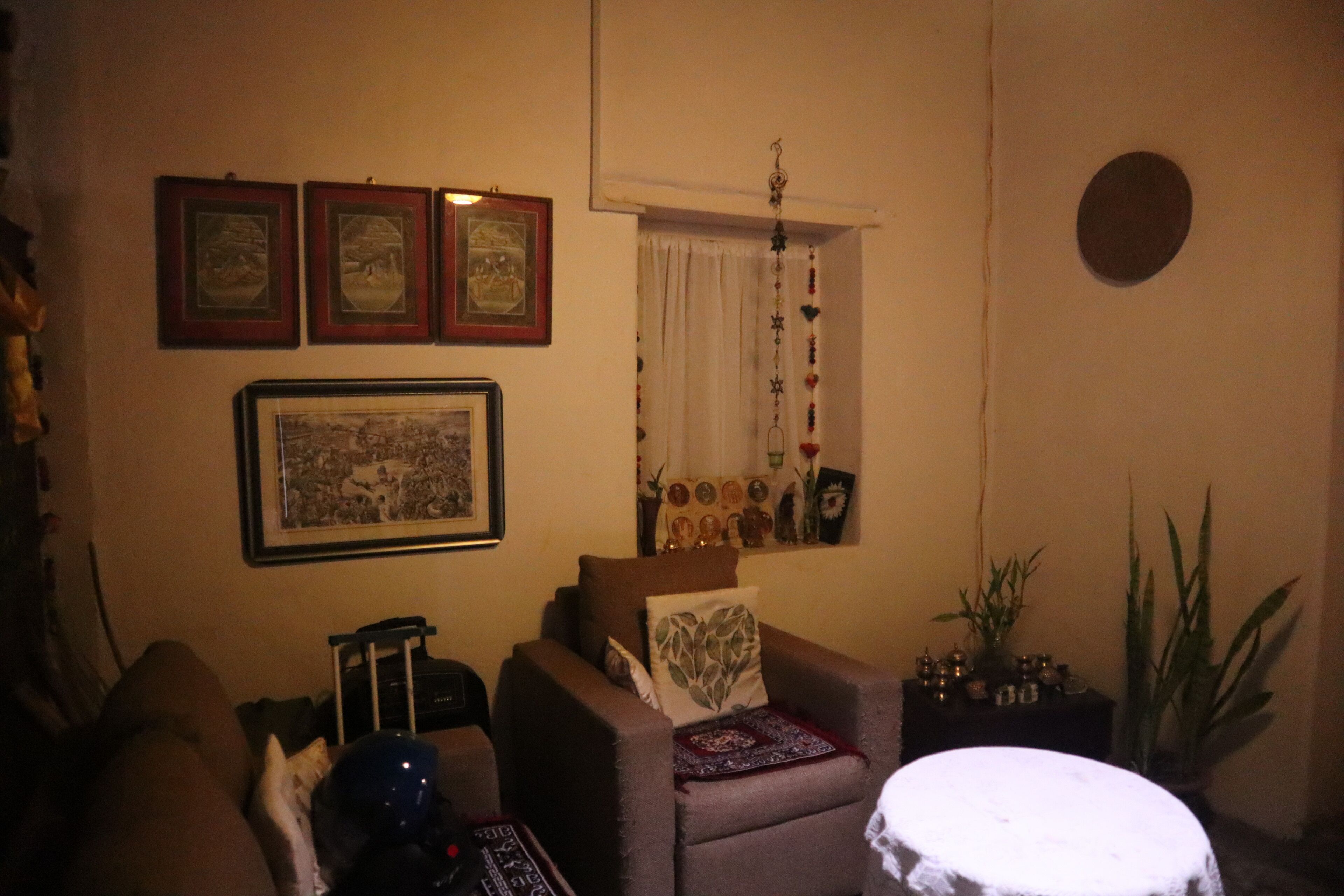 A cozy living room with a sofa, armchair, and a small round table. There are framed pictures on the wall, a window with curtains, and a hanging decoration.