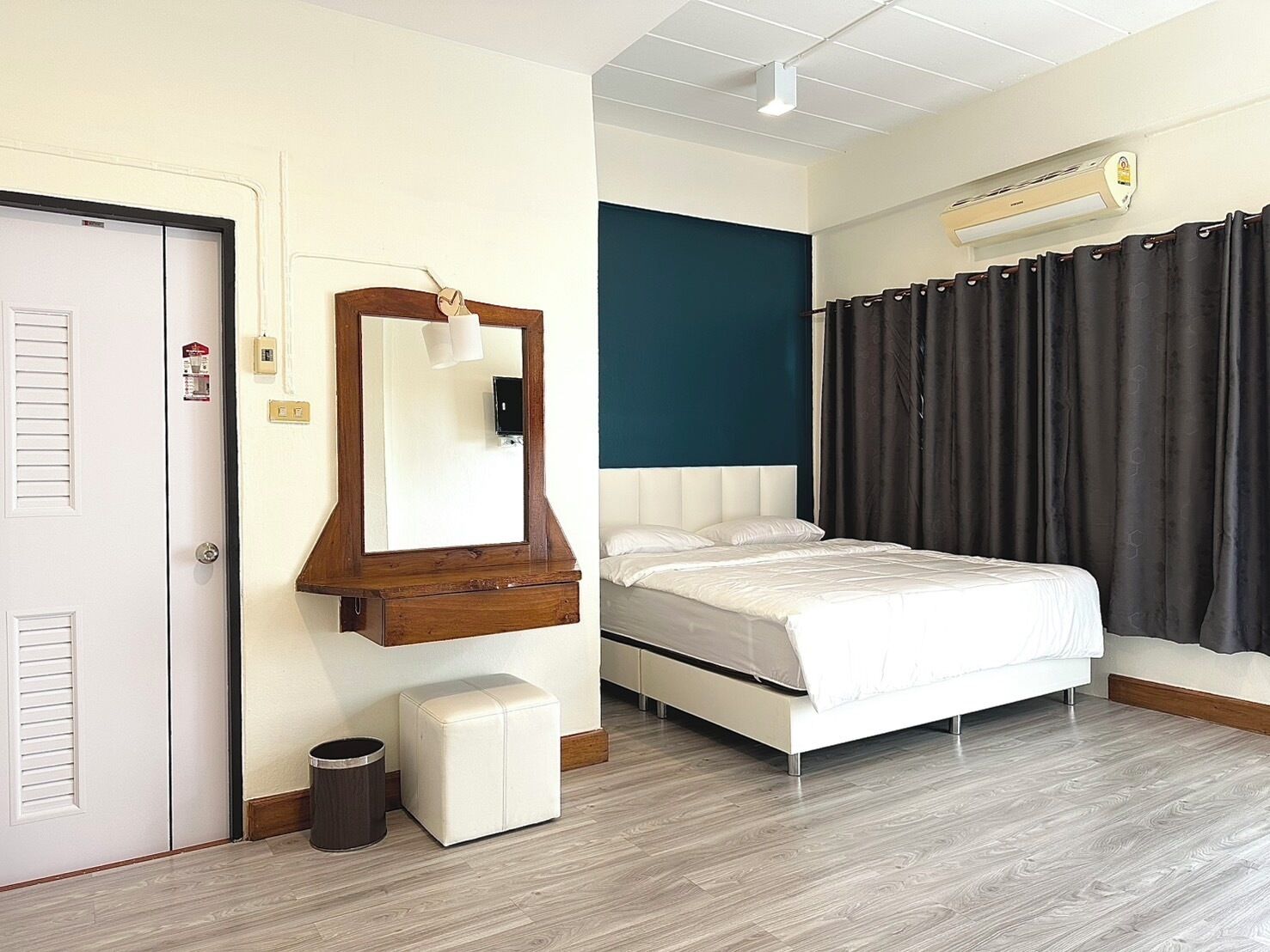 Una camera d'albergo con un letto, una cassettiera in legno, uno specchio, un poggiapiedi bianco e piccolo, un cestino e una finestra con tende.