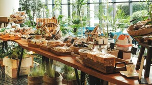 Una mesa de madera llena de diferentes alimentos y cestas, ubicada en una habitación con ventanas amplias y plantas de interior.