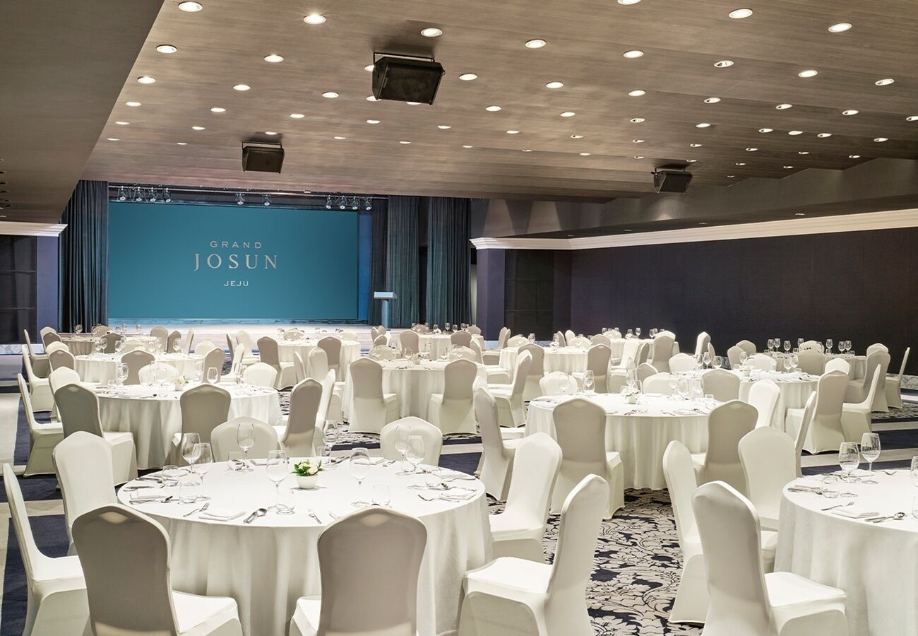A banquet hall with round tables and white chairs, a stage with a blue backdrop, and a modern ceiling design.