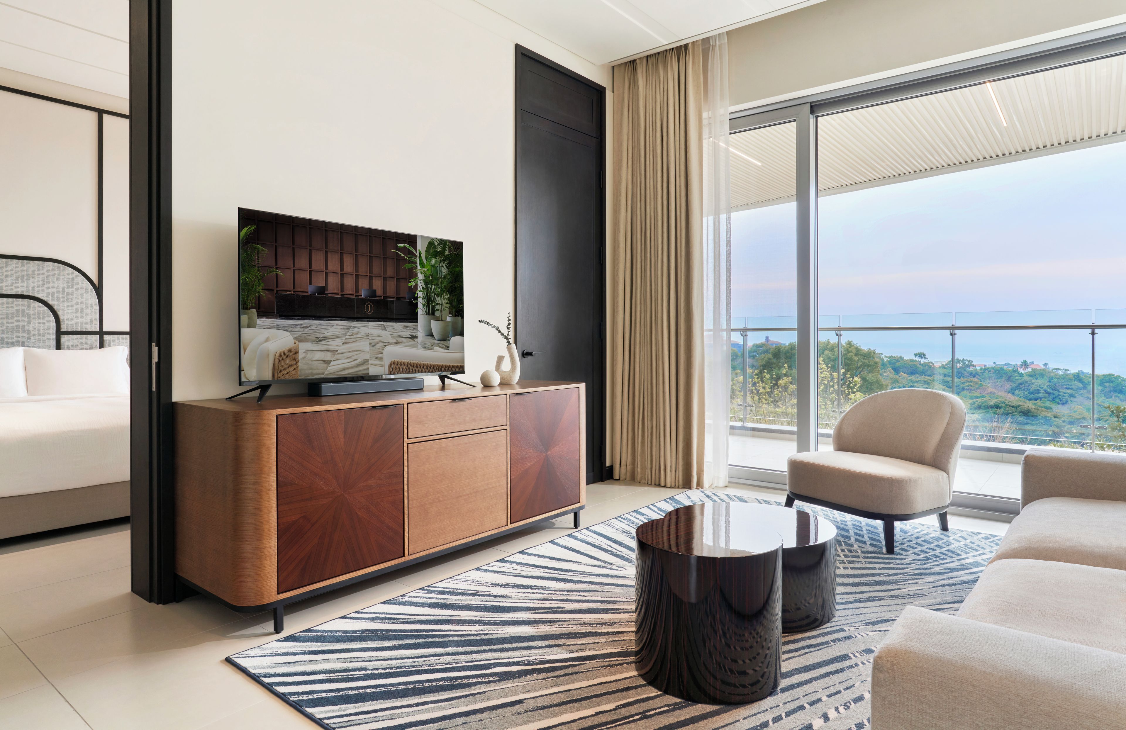 A modern hotel room with a bed, a wooden cabinet, a television, a sofa, a chair, and a large window with a view.