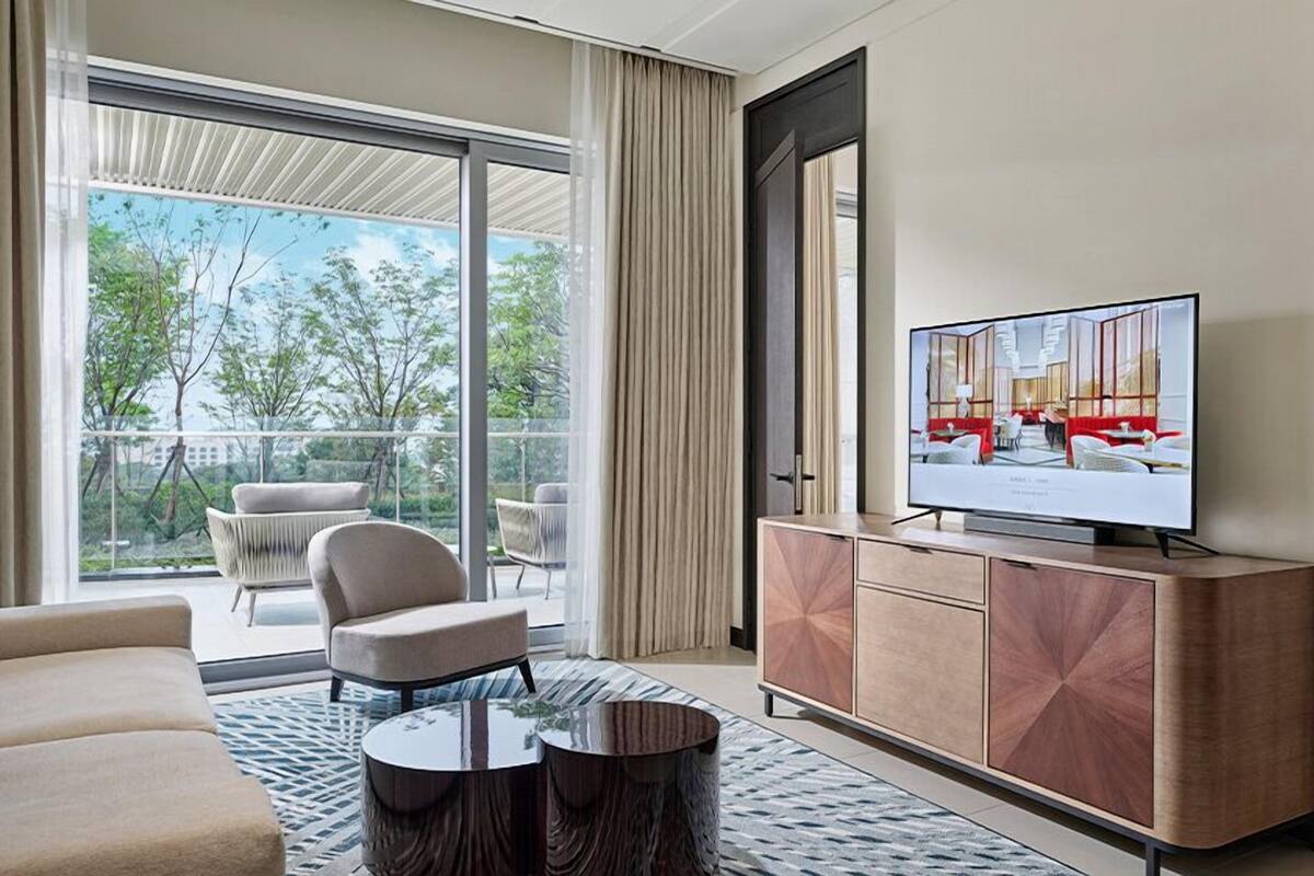 Une chambre d’hôtel moderne dotée d’une grande porte-fenêtre coulissante donnant sur un balcon, d’un téléviseur à écran plat posé sur un meuble en bois et d’un canapé agrémenté d’un tapis à motifs.