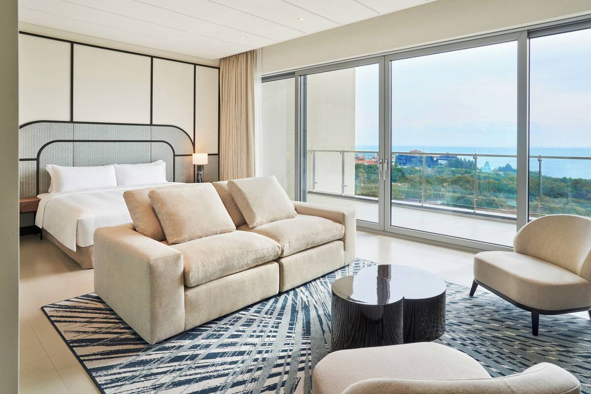 Une chambre d’hôtel moderne dotée d’un grand lit, d’un canapé, d’une table basse et offrant une vue sur la ville grâce à de grandes fenêtres.