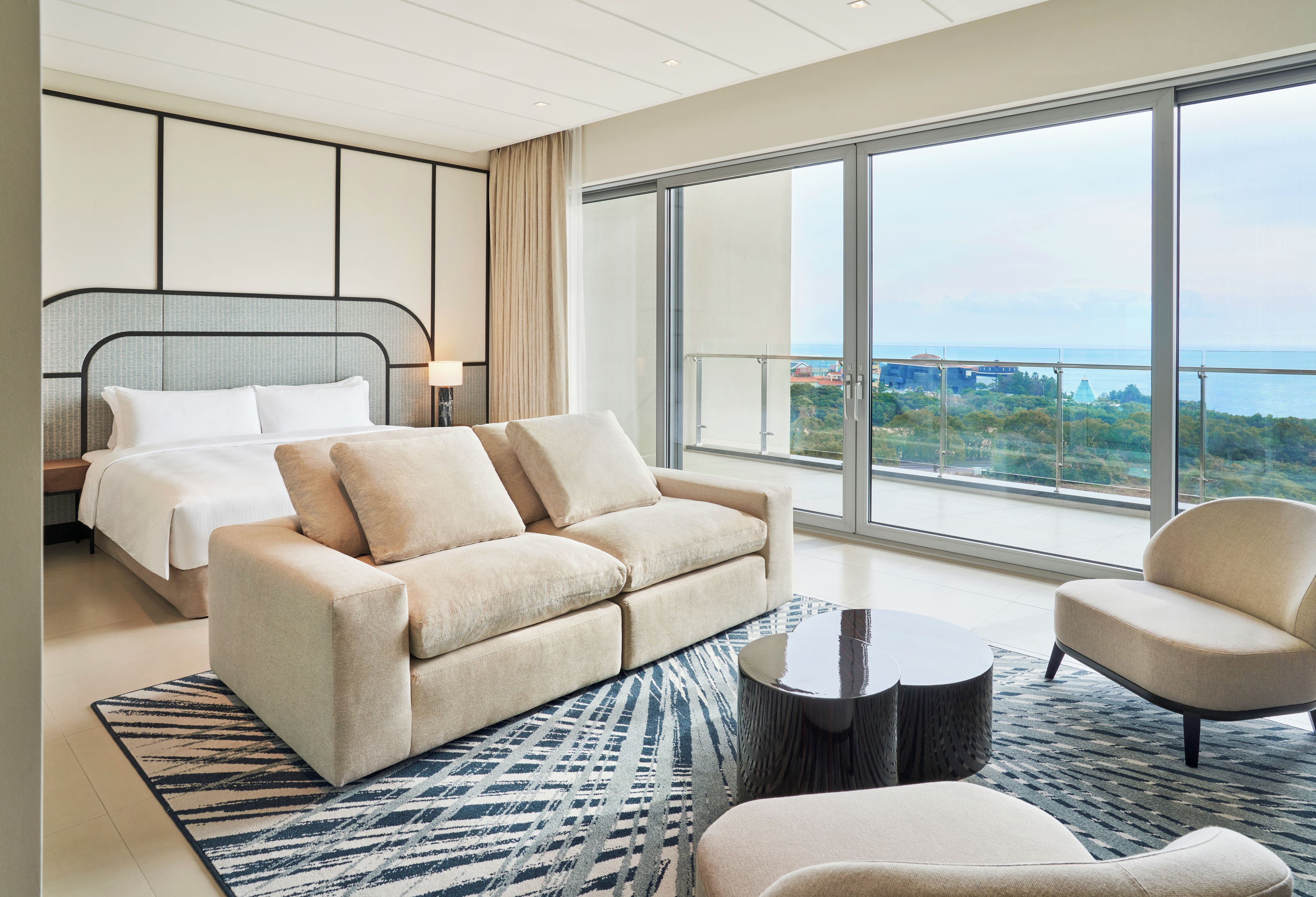 A modern hotel room with a large bed, a sofa, a coffee table, and a view of the city through large windows.