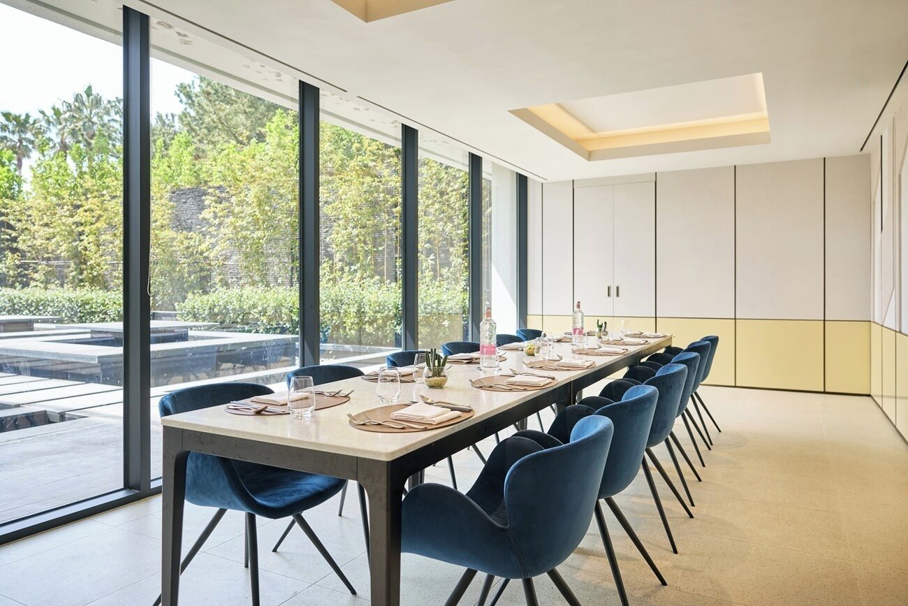 A conference room with a long table, blue chairs, and large windows offering a view of greenery outside.