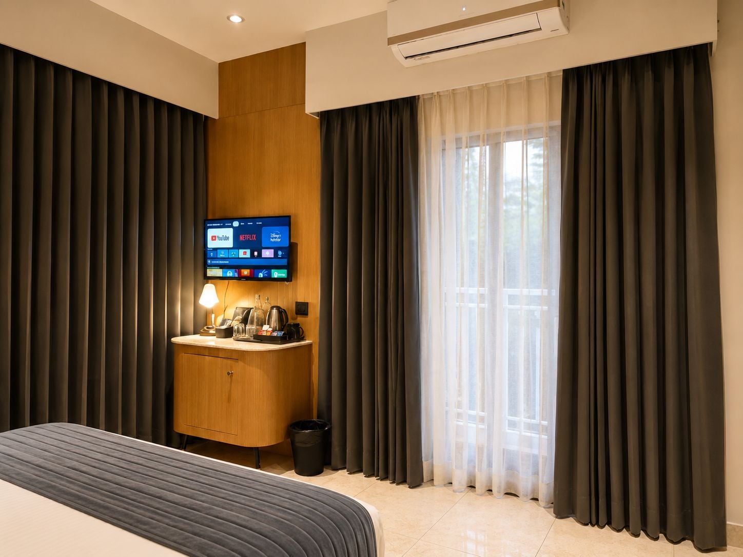Una habitación de hotel con una cama, un escritorio con lámpara, un televisor mostrando servicios de transmisión y una ventana con cortinas.