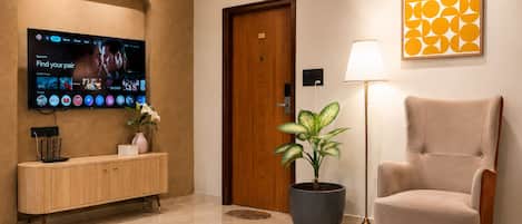 A hotel lobby with a flat-screen TV, a wooden console, a potted plant, a lamp, and a framed artwork.