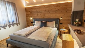 A modern bedroom with a wooden headboard, a grey sofa, a wooden nightstand, and a small wooden table with books and a vase of flowers.