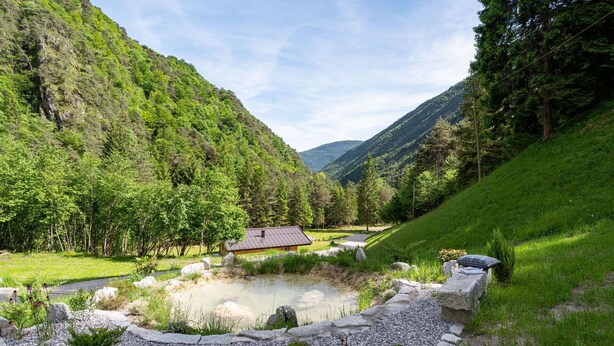 Une maison avec un toit en pente, entourée d’un paysage naturel comprenant un étang et un sentier en pierre.