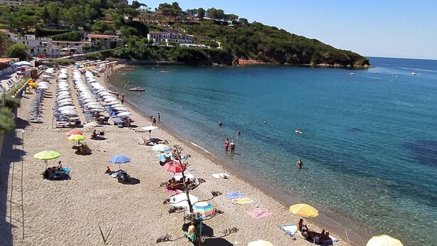 Una spiaggia con file di ombrelloni e lettini, acqua cristallina e palazzi su una collina sullo sfondo.