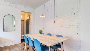 A dining area with a wooden table, blue chairs, a hanging light fixture, and a white brick wall. - Le Plateau Apartment & Suite Near Mont-Royal Ave (Montréal)