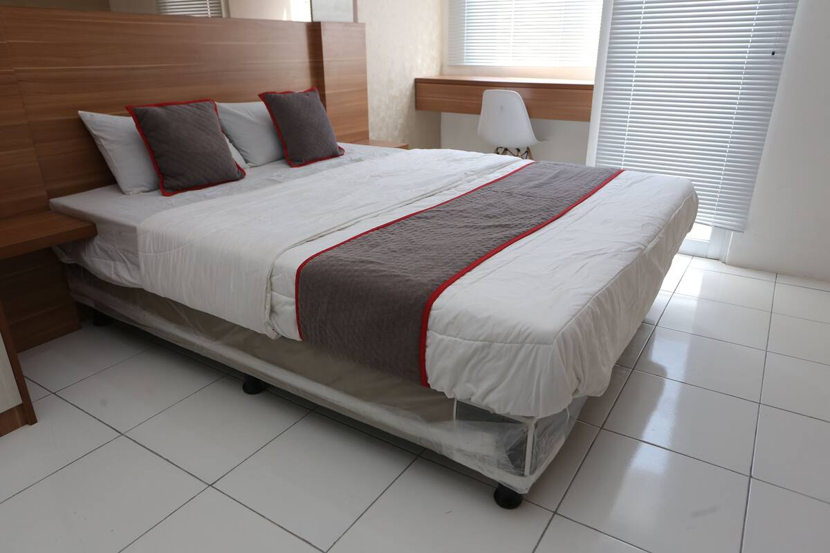Een keurig opgemaakt bed met een houten hoofdeinde, een grijs-witte dekbedovertrek en een rode bies.