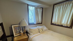 Ein ordentlich bezogenes Bett mit weißer Decke und Kissen, ein Nachttisch aus Holz mit einer Lampe und einem gerahmten Bild, sowie ein Fenster mit Vorhängen.