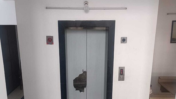 Egy lift, amelyben található egy kezelőpanel, egy láthatóan sérült ajtó és egy kapaszkodó.
