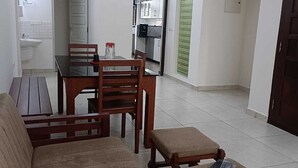 Egy nappali, amelyben van egy kanapé, egy dohányasztal és egy étkezőasztal székekkel. A mennyezeten egy ventilátor található, egy nyitott ajtón keresztül belátható a konyha, és egy zöld színű ajtó is van.