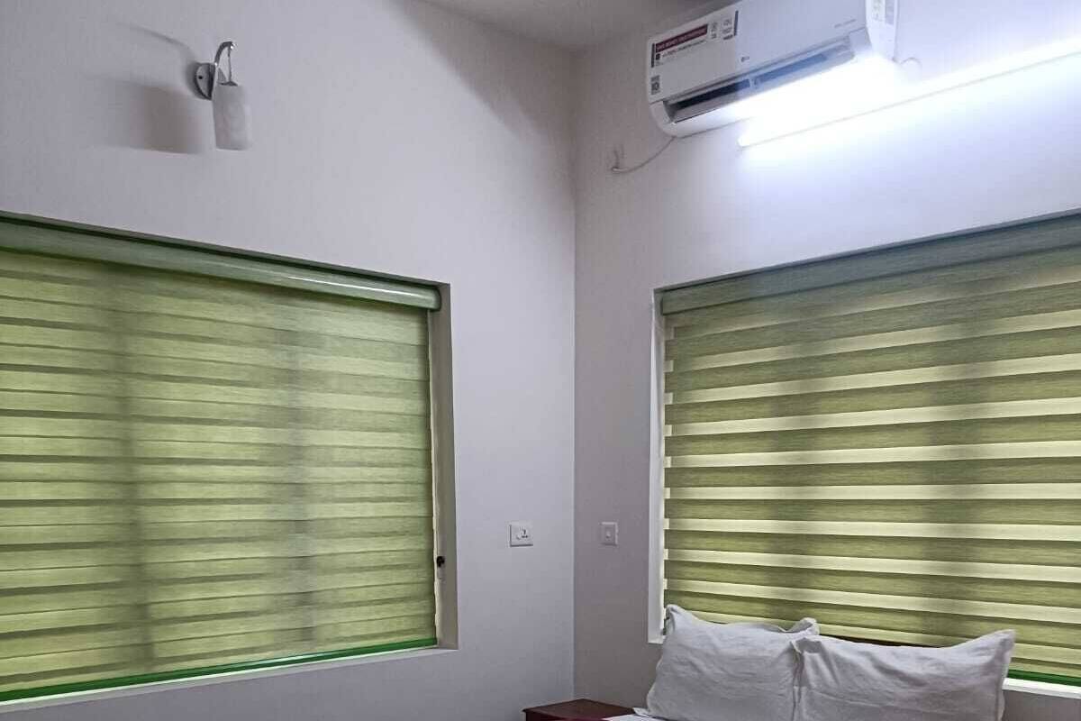 A bedroom with a bed, a bedside table, a window with blinds, an air conditioning unit, and a wall-mounted light fixture.