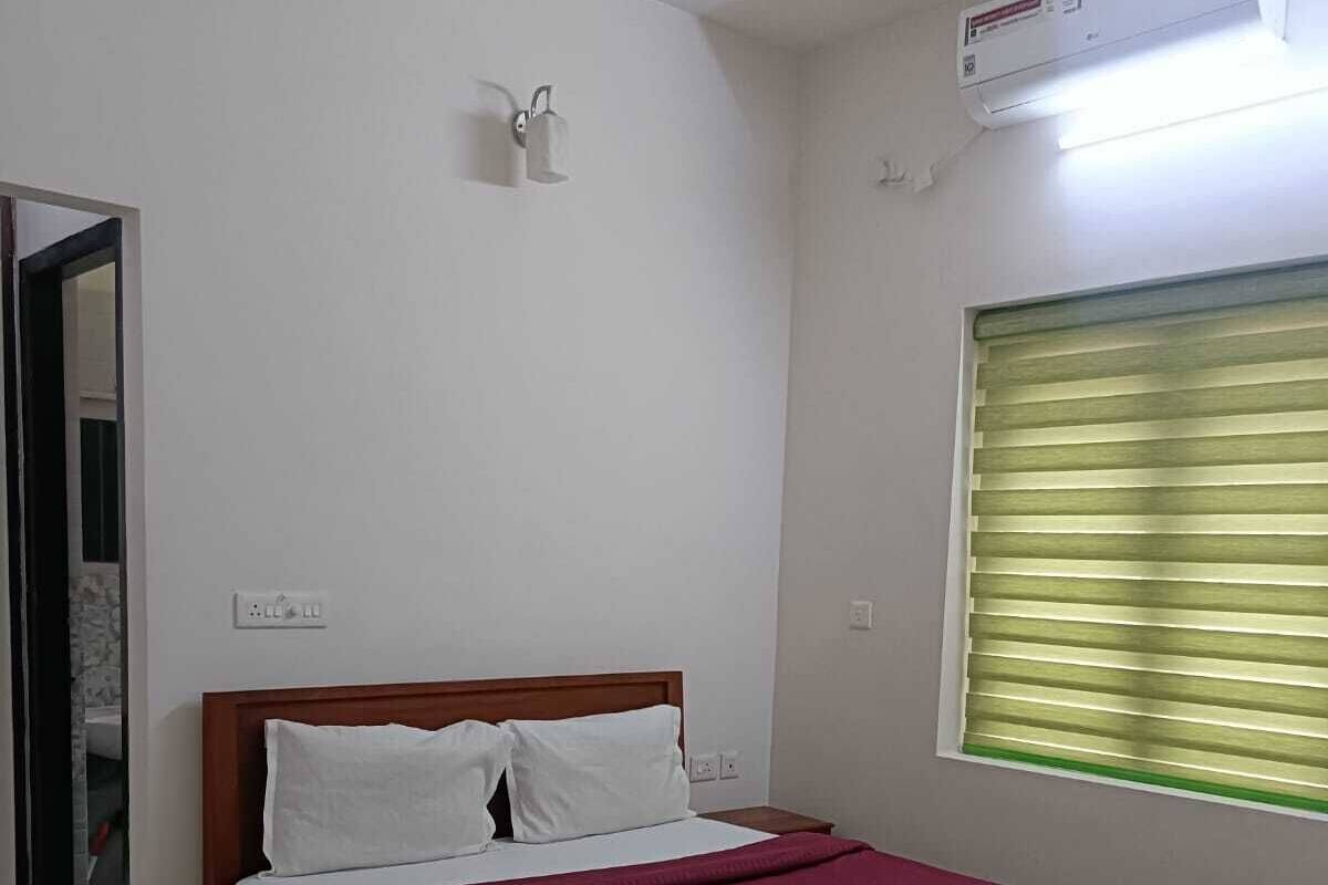 A bedroom with a wooden bed, white bedding, a window with blinds, an air conditioning unit, and a small bedside table.