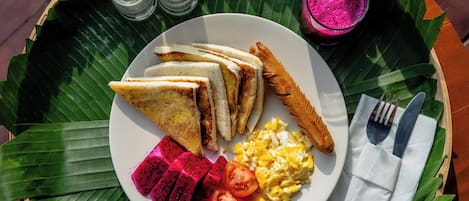A plate of food including toast, eggs, and fruit, served on a banana leaf with a fork and knife on a wooden table.