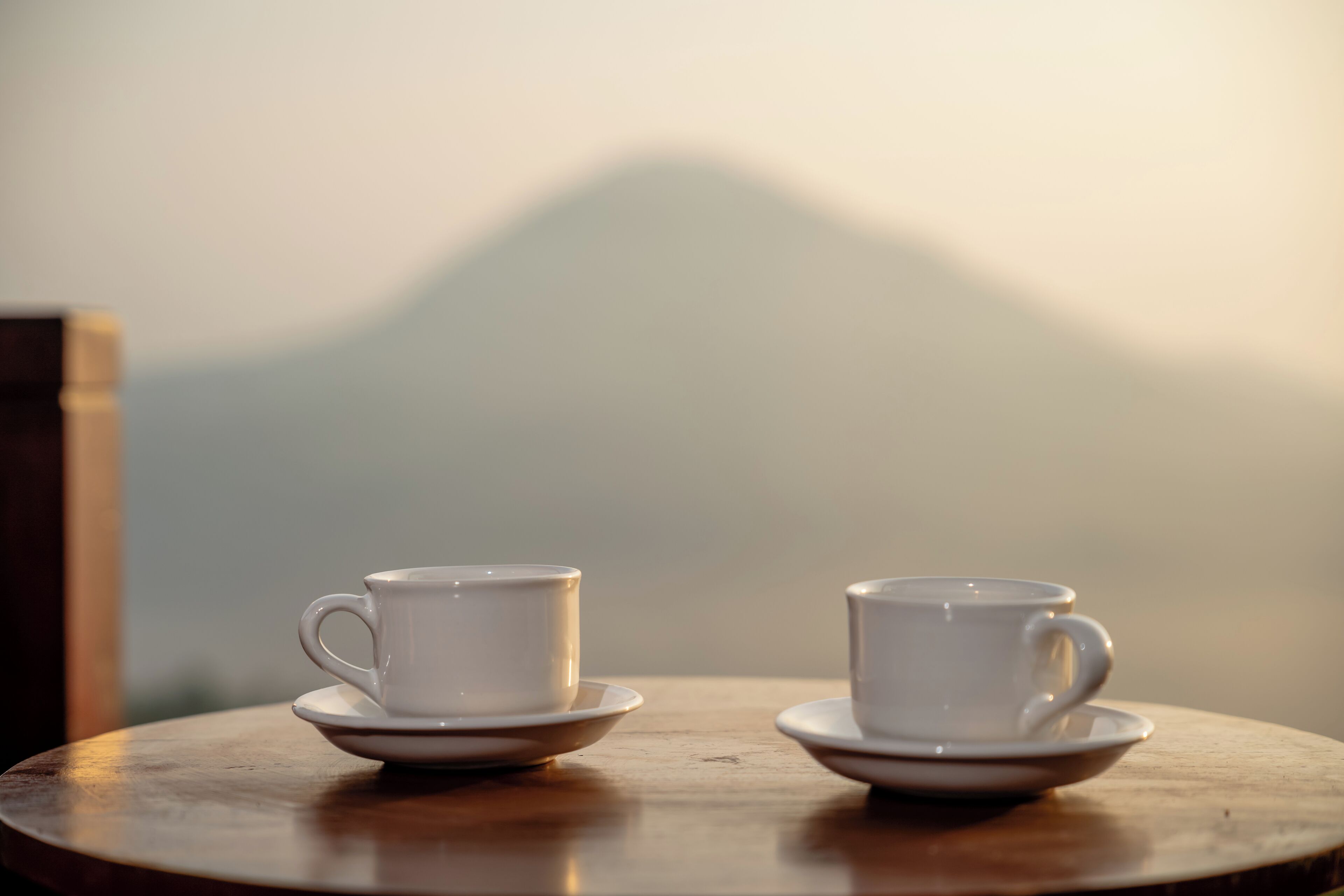 Two white cups on saucers on a round wooden table with a blurred mountain view in the background.