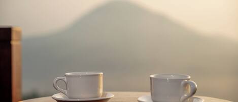 Two white cups on saucers on a round wooden table with a blurred mountain view in the background.