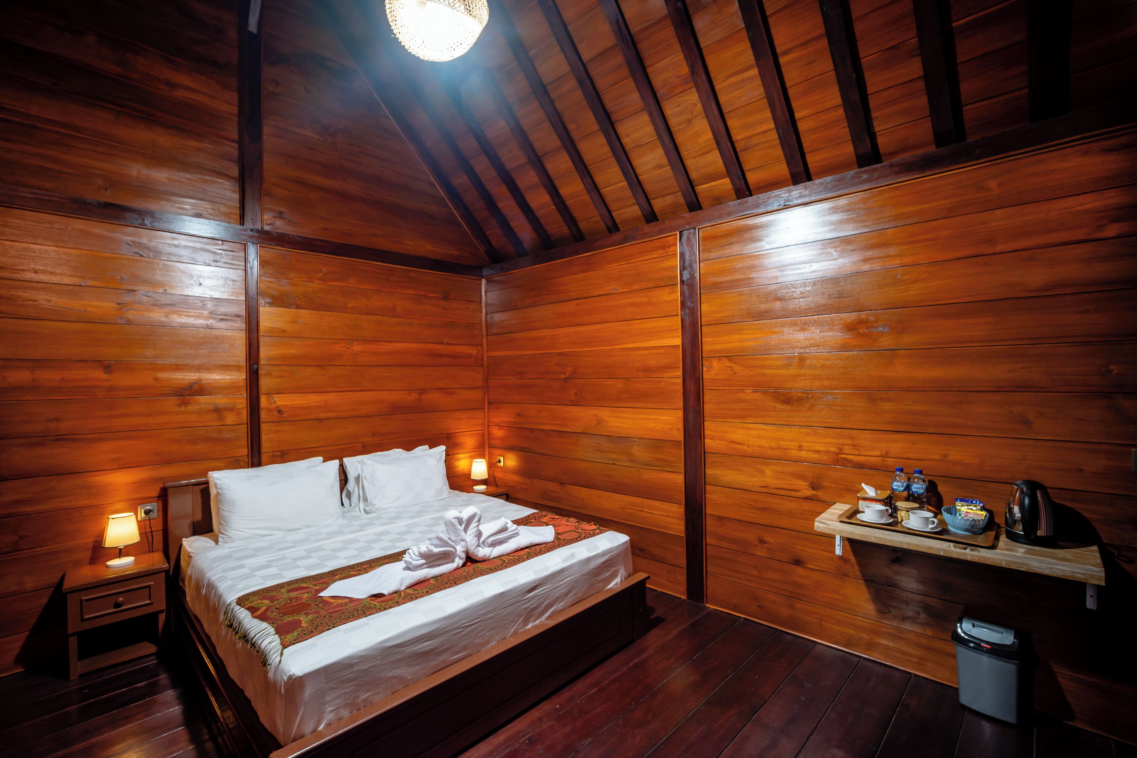 A wooden-paneled room with a bed, bedside lamps, a shelf with toiletries, and a trash bin.