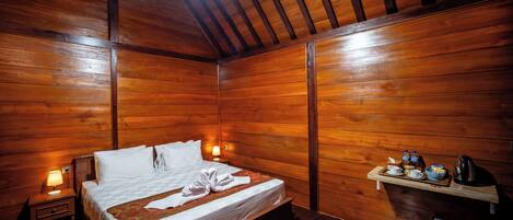A wooden-paneled room with a bed, bedside lamps, a shelf with toiletries, and a trash bin.