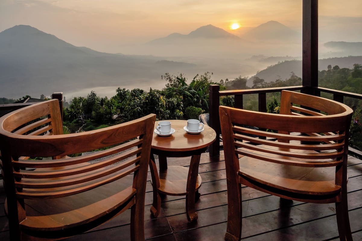 A wooden deck with two chairs and a table, overlooking a mountainous landscape with a sunrise or sunset.