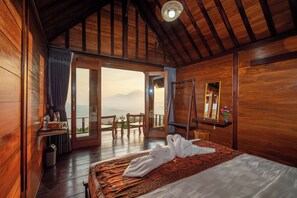 A wooden room with a bed, a table with a bottle and a cup, a chair, and a view of mountains through an open door. - Pondok Devana Kintamani (Kintamani)