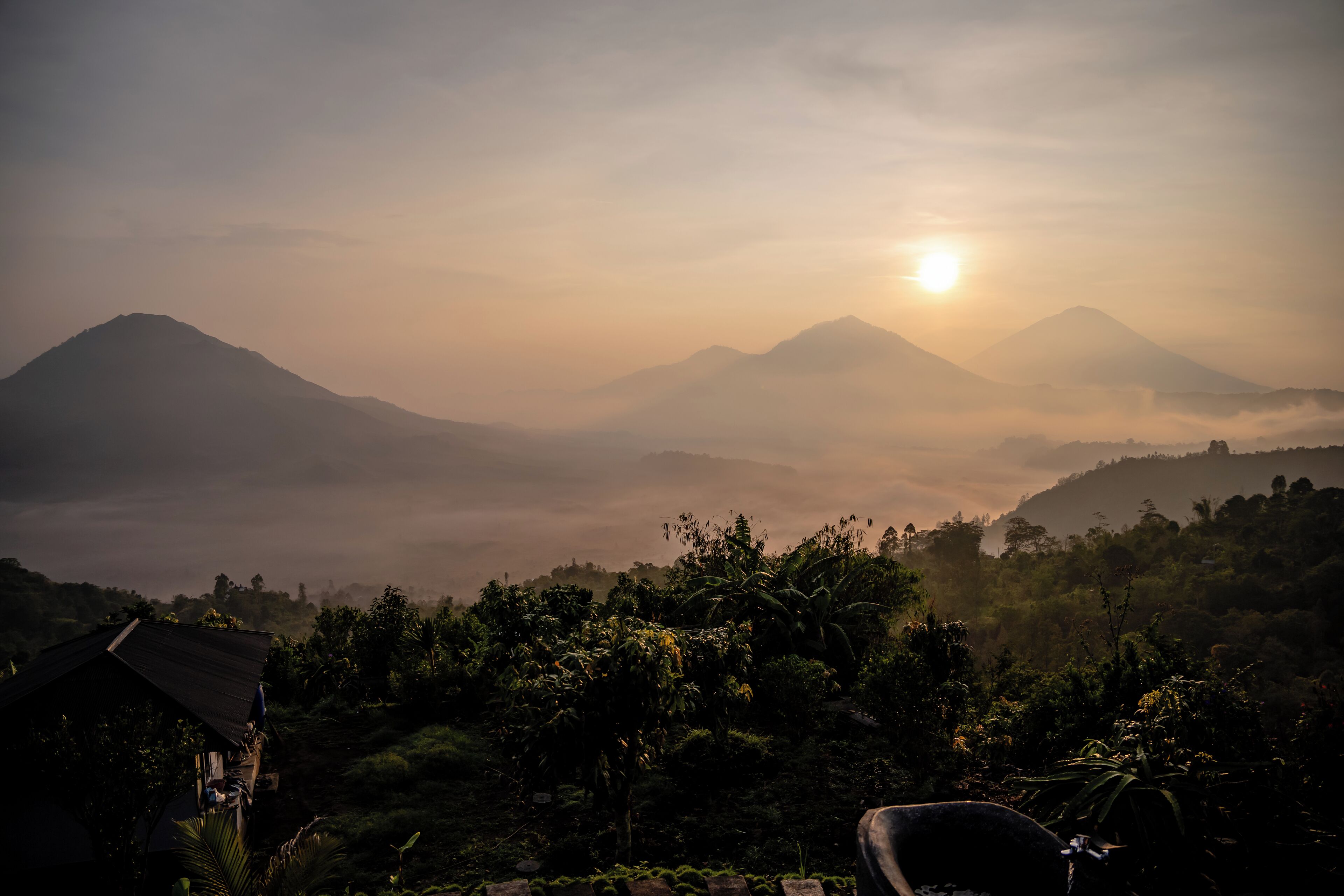 A mountainous landscape with a sunrise or sunset, a house with a sloped roof, and dense vegetation.