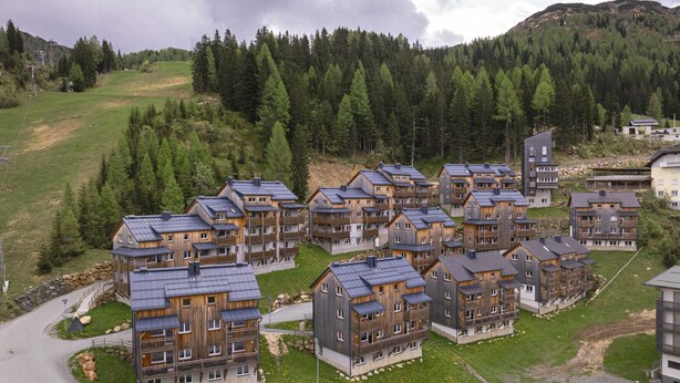 Un gruppo di edifici moderni a più piani, con facciate in legno e tetti scuri, immerso in un paesaggio verdeggiante e una fitta foresta.