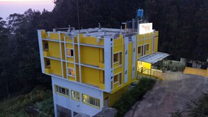A multi-story building with a yellow and white color scheme, featuring a rooftop water tank and a sign with a bird logo. - VS Cube Estancia (Yercaud)