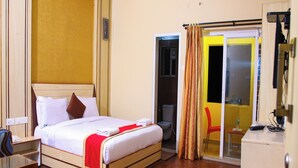 A hotel room with a bed, a desk, a chair, a small table, and a bathroom visible through a glass door. - VS Cube Estancia (Yercaud)