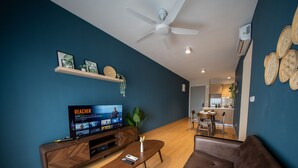 A modern living room with a flat-screen TV, wooden furniture, a ceiling fan, and a kitchen in the background. - Maya Bay Residences (Banting)