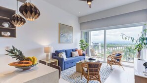 A modern living room with a blue sofa, a dining area with wicker chairs, a large window with a city view, and a ceiling fan.