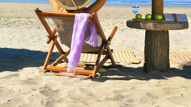 A beach scene with a wooden chair under a thatched umbrella, a table with apples and a heart-shaped balloon, and the ocean in the background. - El Tigre Luxe & Cozy Stay: Pool, Balcony and WiFi! (Nuevo Vallarta)