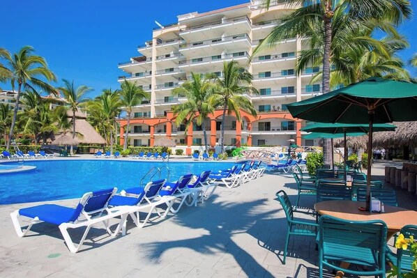A resort pool area with blue lounge chairs, a swimming pool, palm trees, and a multi-story building with balconies.