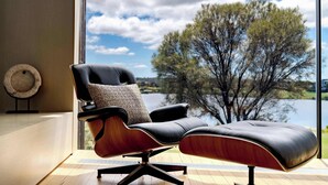 A modern lounge chair with a cushioned seat and backrest, positioned by a large window overlooking a lake and trees. - Front Row Golf & Lake Views Luxe Coastal Retreat (Torquay)