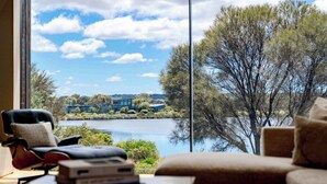 A room with a large window overlooking a lake and buildings, a chair, a table with books, and a sofa. - Front Row Golf & Lake Views Luxe Coastal Retreat (Torquay)