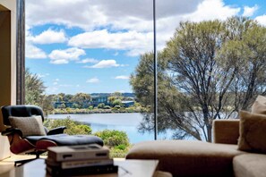 A room with a large window overlooking a lake and buildings, a chair, a table with books, and a sofa.